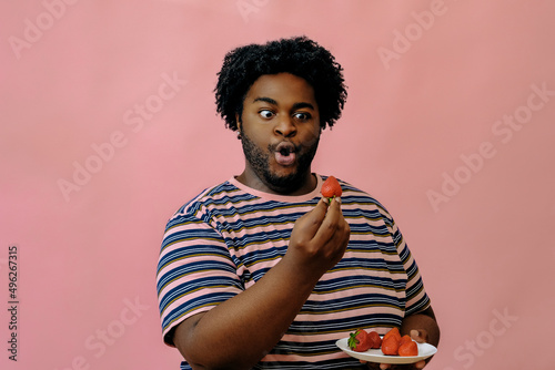young happy african american man eating strawberries in the studio over pink background