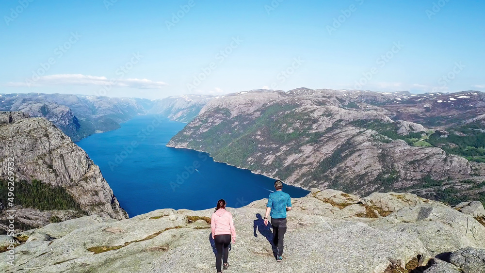 Naklejka premium Couple sitting on a rock and admiring a view on Lysefjorden. Areal shot, upper perspective on the couple. Endless view of the fjord. Couple rising hands above their head and forming a heart. Freedom