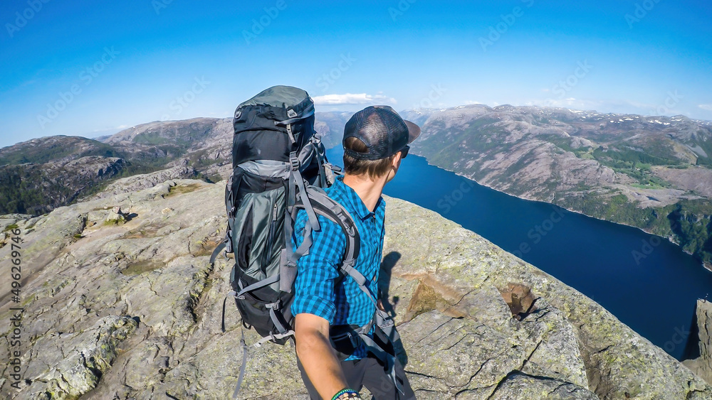 A young man in a hiking outfit, with a huge hiking backpack walks along the edge of a steep cliff , with a selfie stick. In the back is Lysefjorden, Norway. He walks on a barren rocky cliff