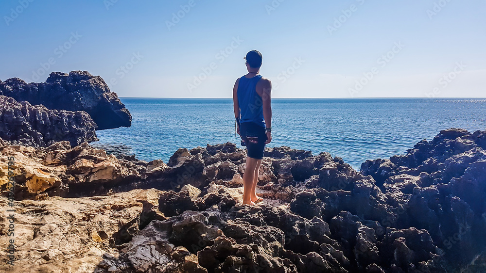 Young man standing in between the steep cliffs of Cape Greco, Cyprus. The cliff looks dangerous. Barren slopes of the stony formation. Sea has many shades of blue. Endless line of a horizon. Clear day
