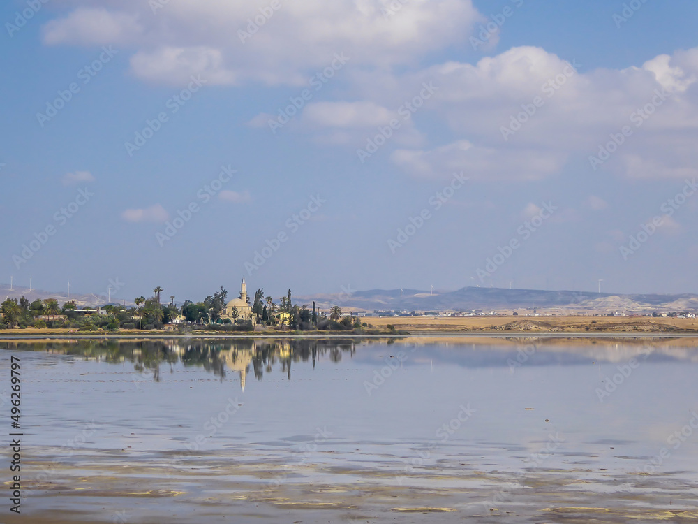 Hala Sultan Tekke seen from a distance. The mosque is surrounded by lush setting - palm trees and smaller bushes. Clear reflection of the mosque in the calm surface of a Larnaca Salt Lake.