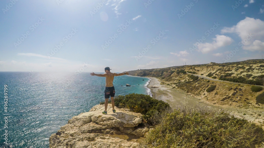 A young man standing at the steep cliff and spreading his arms wide in a gesture of freedom in Paphos, Cyprus. Blue sea spreads in front of him. Barren slopes of the cliff. Endless horizon line.