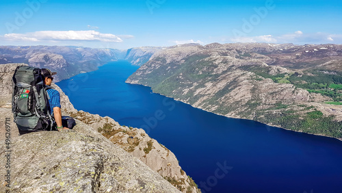 A man wearing blue shirt, full cap and a huge hiking backpack sits at the edge of a steep cliff of Preikestolen. A view on Lysefjorden. Fjord goes far inland. Man enjoys the view, feels free and happy © Chris