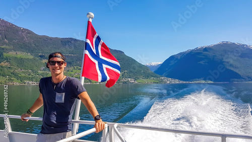 A young man leaning on a rail of a ship with a Norwegian flag waving behind him, Songefjorden, Norway. The motor of the ship makes the water wavy and foamy. Lush green mountains surrounding the fjord.