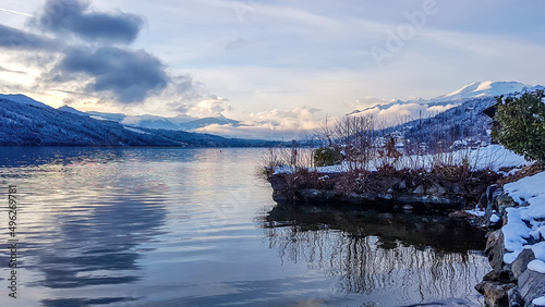 A beautiful view on a Millstaetter lake in Austria. The lake is surrounded by Alps. Mountains are covered with snow. Soft colors of the sunset. Blue sky. The shore is overgrown with bushes. photo