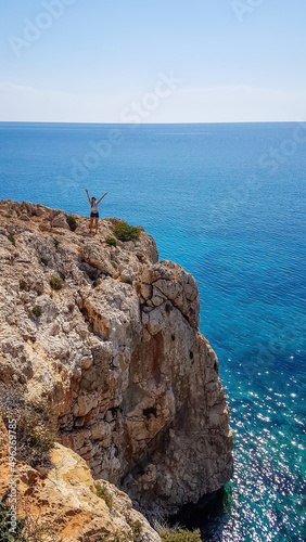 Girl standing at the steep cliff of Cape Greco, Cyprus, spreads her hands wide. The cliff looks dangerous. Barren slopes of the stony formation. Sea has many shades of blue. Endless line of a horizon photo
