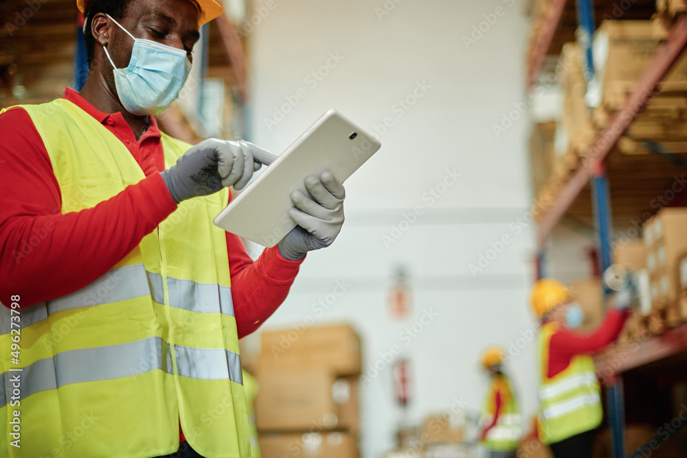 Afro male worker using a tablet checking the transport of a box in a warehouse wearing a safety mask and work waistcoat. focus on tablet Afro male worker using a tablet checking the transport of a box in a warehouse wearing a safety mask and work waistcoat. focus on tablet