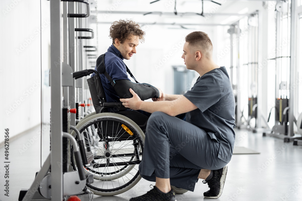 Rehabilitation specialist helps a guy in a wheelchair to do exercises for recovery from injury. Concept of physical therapy for people with disabilities Rehabilitation specialist helps a guy in a wheelchair to do exercises for recovery from injury. Concept of physical therapy for people with disabilities