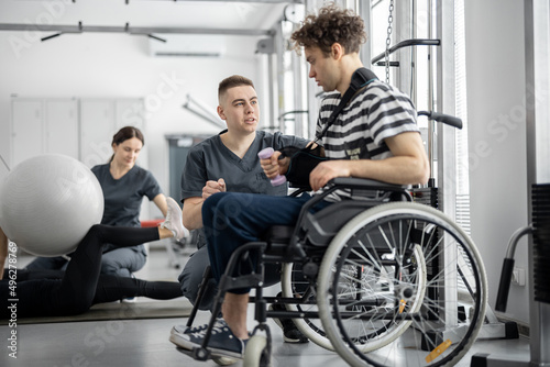 People doing exercises at rehabilitation center. Worker helps a guy in a wheelchair to do exercises for recovery from injury. Concept of physical therapy for people with disabilities