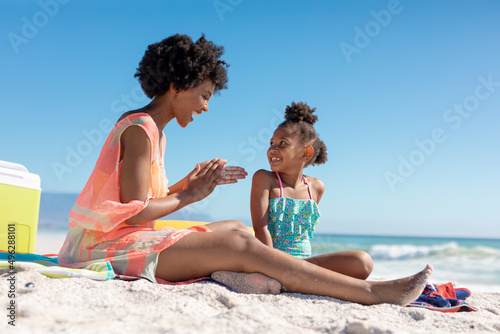 Happy african american woman applying suntan lotion on daughter while sitting at beach on sunny day
