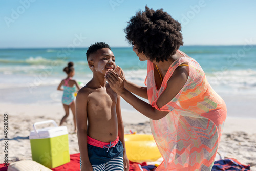 African american woman applying suntan lotion on son's cheeks at beach during sunny day