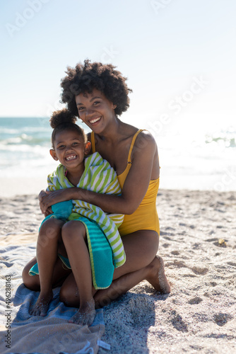 Portrait of happy african american mother and daughter sitting on sand at beach during sunny day