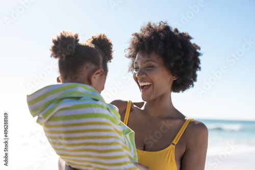 Cheerful african american mother with daughter wrapped in towel at beach on sunny day