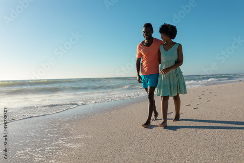 Happy african american couple walking together at beach against clear blue sky on sunny day