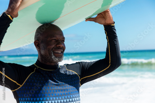 Canvas Print Smiling african american bald retired senior man carrying surfboard on head at b