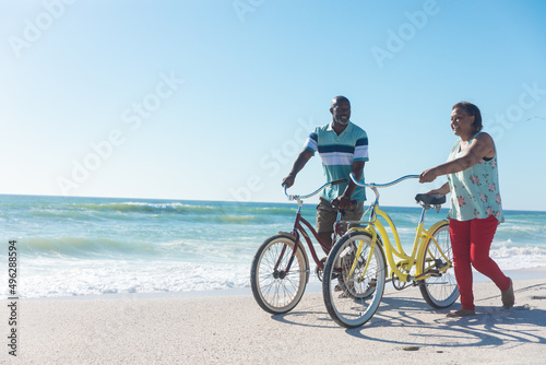 African american senior couple wheeling bicycles at beach against blue sky with copy space