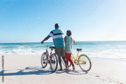 Rear view of african american senior couple wheeling bicycles at beach with copy space on blue sky
