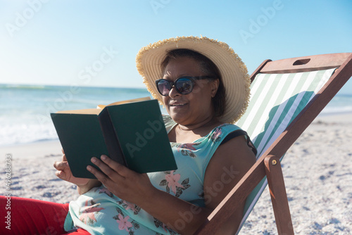 African american retired senior woman reading book while sitting on folding chair at beach