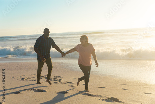 Active african american senior couple holding hands running on shore at beach during sunset