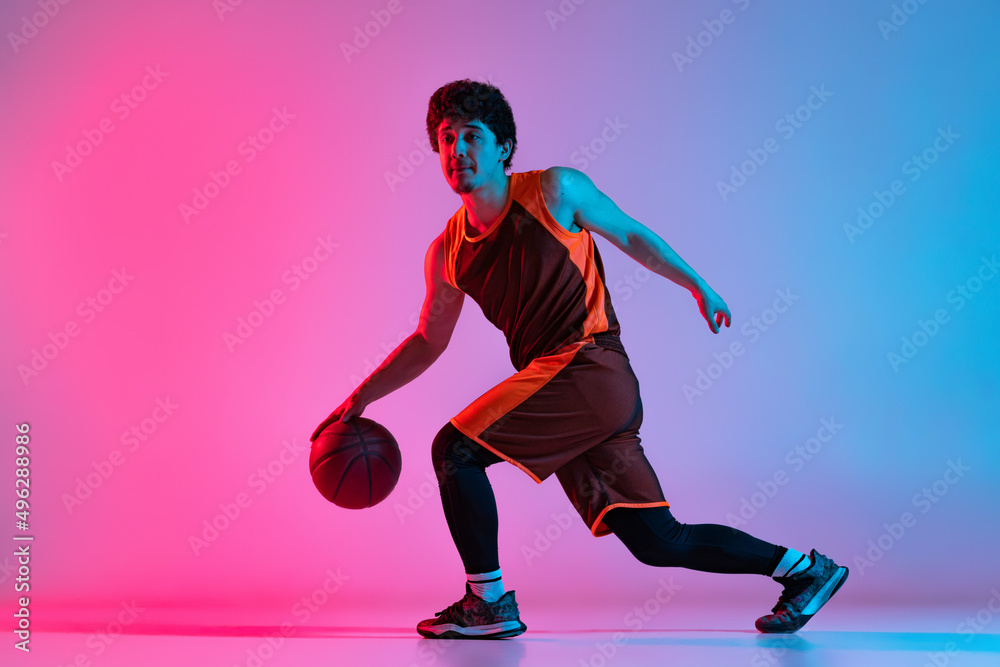 Sportive man playing basketball isolated on gradient pink blue studio background in neon light. Youth, hobby, motion, activity, sport concepts.
