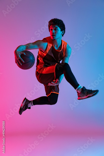 Young energetic man playing basketball isolated on gradient pink blue studio background in neon light. Youth, hobby, motion, activity, sport concepts.