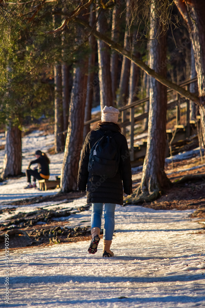 The Walking Woman and the Resting Athlete. Asian woman walking through a snow-covered path in the forest towards a resting athlete woman during the end of the winter season on a sunny afternoon.