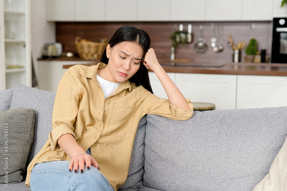 Unhappy young asian woman sitting on sofa, covered forehead with hand and feel frustration, tired and depressed eastern female alone at home, suffering from headache, feels unwell Unhappy young asian woman sitting on sofa, covered forehead with hand and feel frustration, tired and depressed eastern female alone at home, suffering from headache, feels unwell
