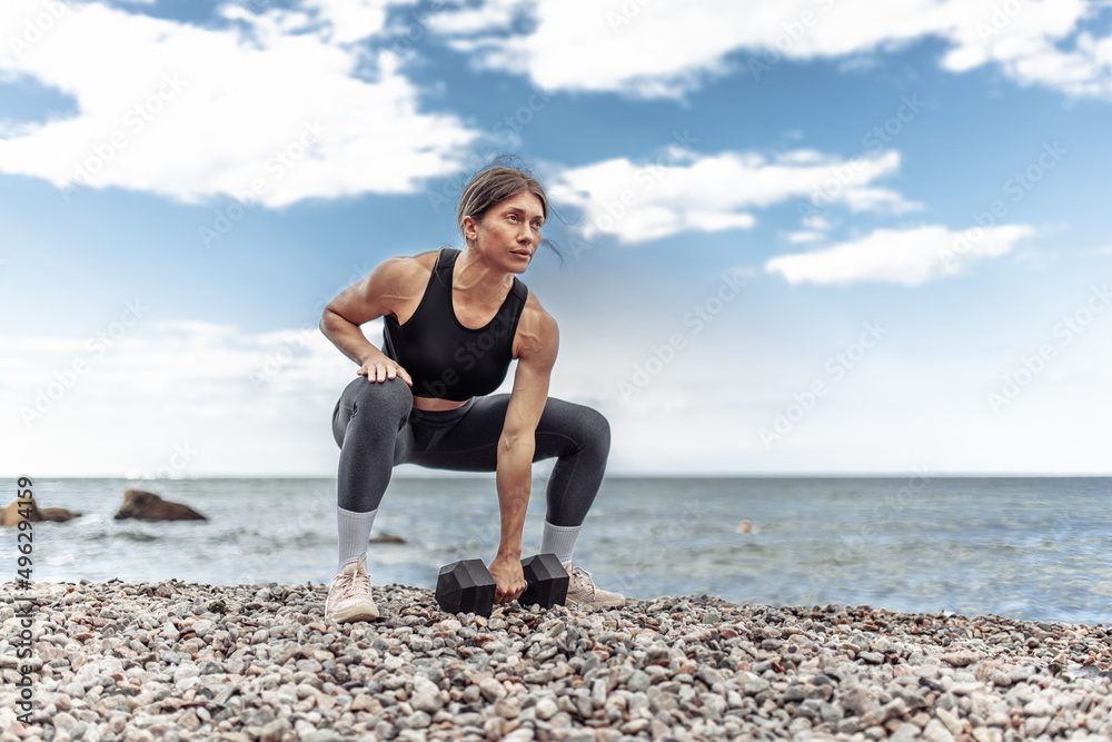 Strong athletic woman exercising with heavy dumbbell on the beach during the day with blue sky and clouds. Functional outdoor training