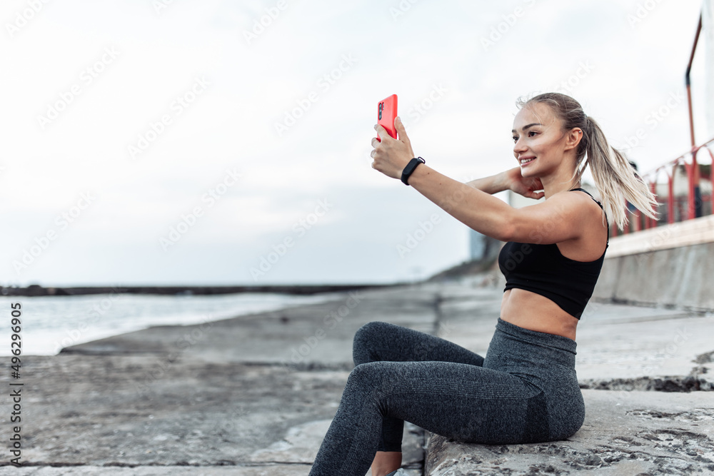 Fit woman in sportswear makes selfie on phone while sitting on urban beach