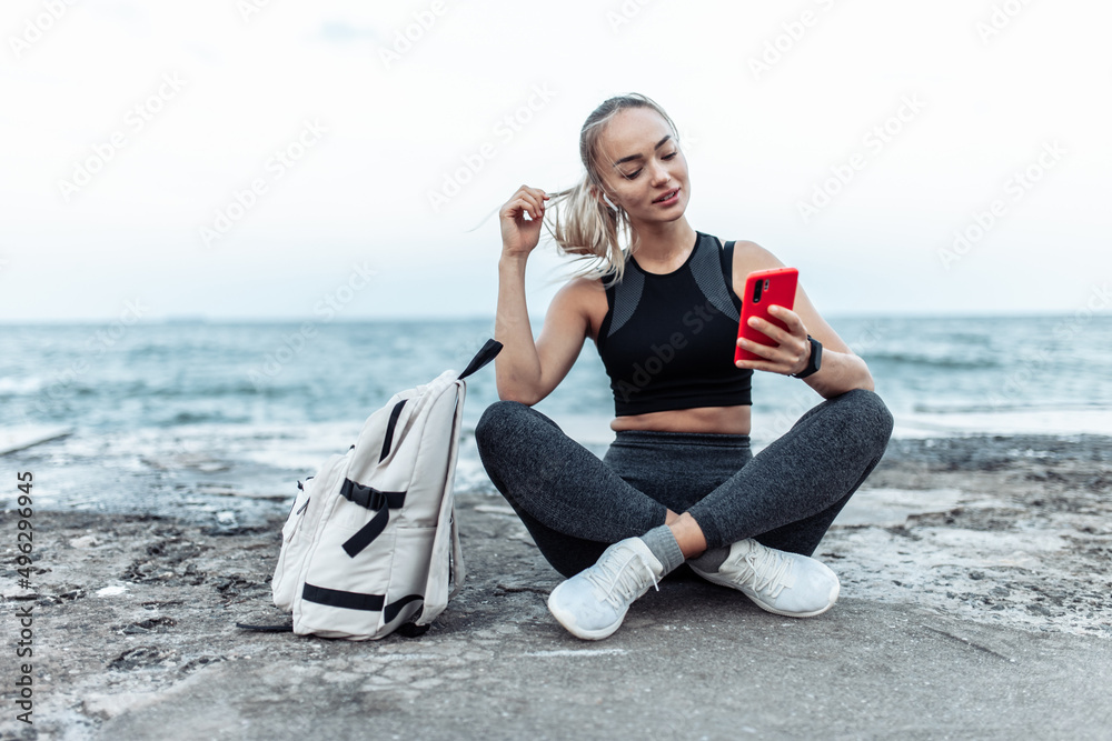 Happy Fit woman in sportswear using smartphone and listen music in headphones while sitting on urban beach