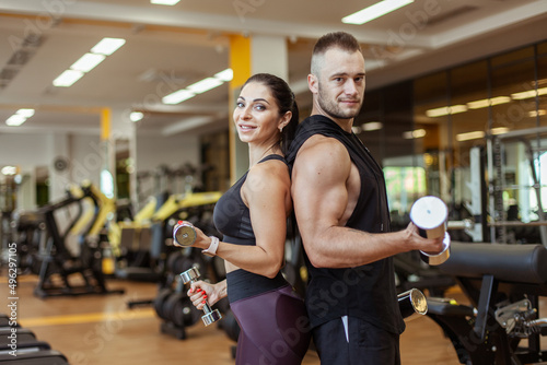 Young athletic man and woman standing back to back, holding dumbbells in hands, looking at camera in the gym