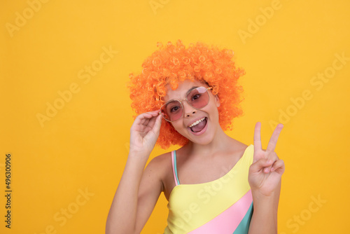 happy kid in sunglasses and swimsuit wearing orange curly wig hair showing peace gesture, peace