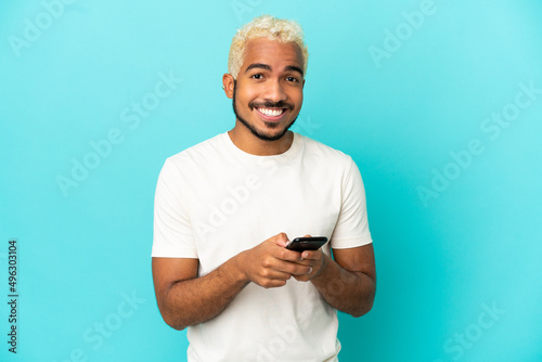 Young Colombian handsome man isolated on blue background sending a message with the mobile