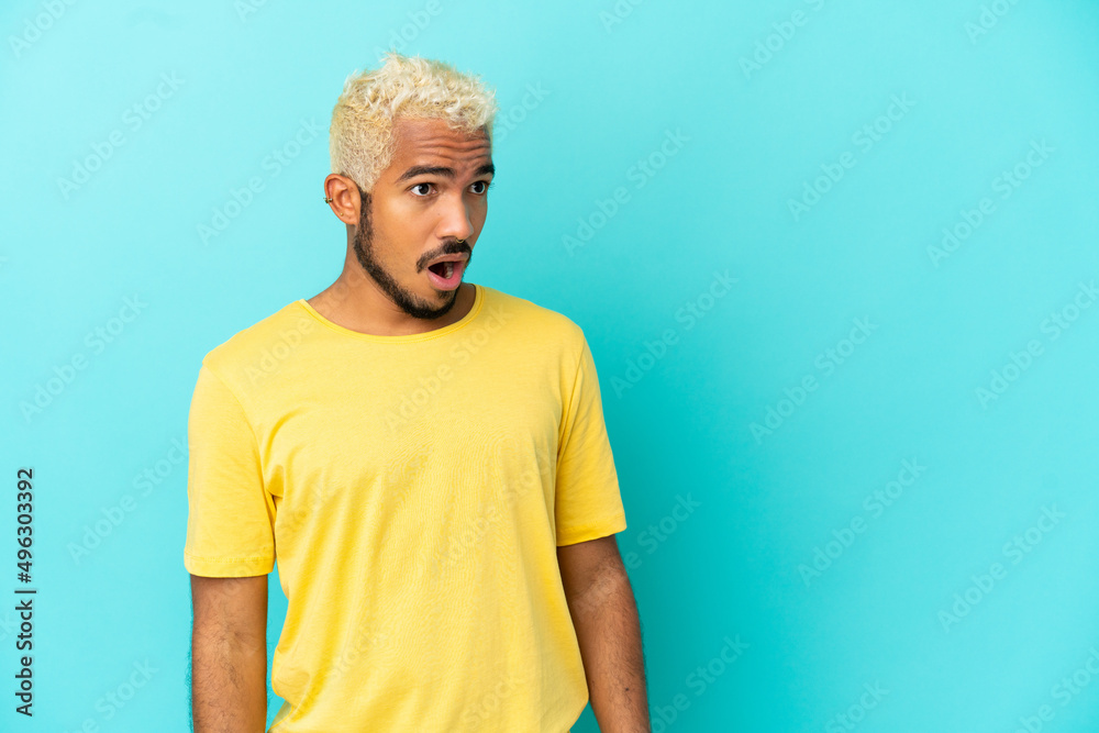 Young Colombian handsome man isolated on blue background doing surprise gesture while looking to the side
