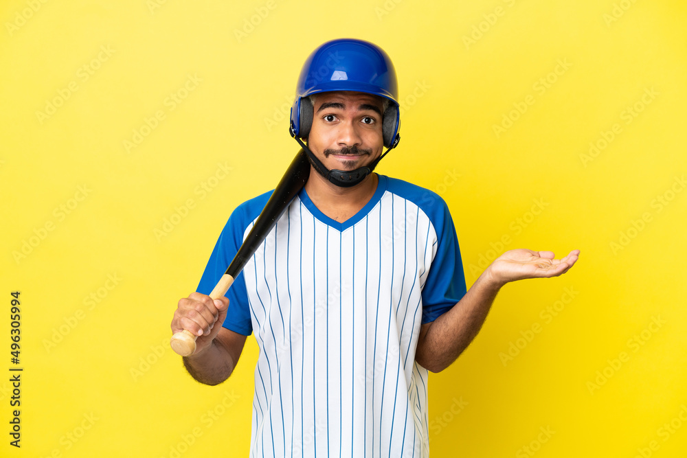 Young Colombian latin man playing baseball isolated on yellow background having doubts while raising hands