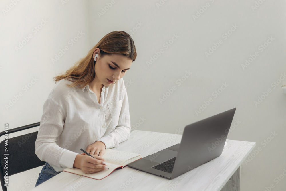 A young beautiful girl in headphones learns with internet chat, a Skype teacher prepares for an exam, a schoolgirl learns online, watches a webinar, takes notes, looks at a laptop, distance learning A young beautiful girl in headphones learns with internet chat, a Skype teacher prepares for an exam, a schoolgirl learns online, watches a webinar, takes notes, looks at a laptop, distance learning