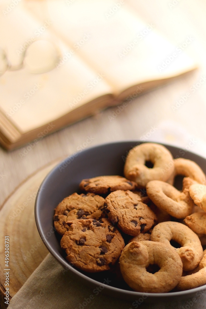 Plate of chocolate chip cookies and sugar cookies, vintage books and reading glasses, candle and flower on the table. Hygge at home. Selective focus.