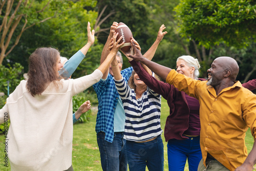 Happy multiracial active senior male and female friends holding rugby ball with hands raised