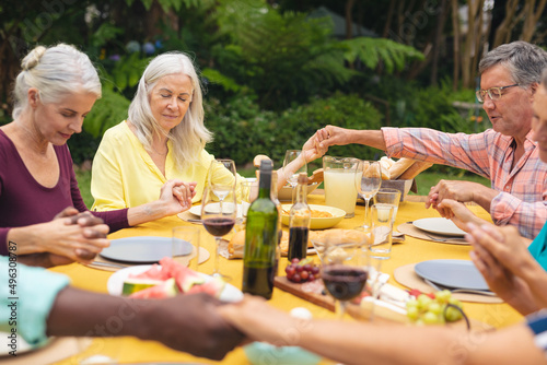 Multiracial senior male and female friends holding hands and praying at table in backyard