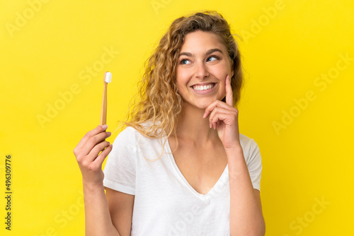 Young caucasian woman holding a brushing teeth isolated on yellow background thinking an idea while looking up