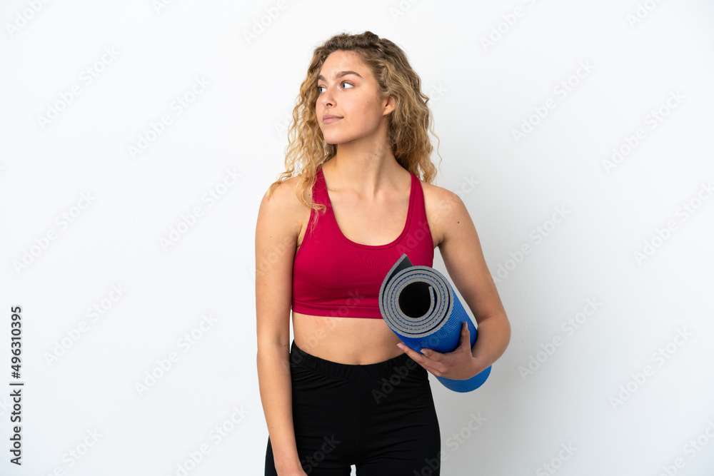 Young sport blonde woman going to yoga classes while holding a mat isolated on white background looking to the side Young sport blonde woman going to yoga classes while holding a mat isolated on white background looking to the side