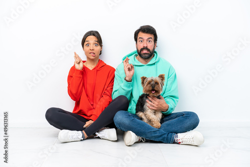 Young caucasian couple sitting on the floor with their pet isolated on white background with fingers crossing
