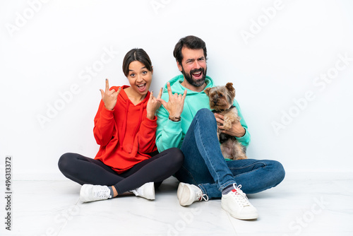 Young caucasian couple sitting on the floor with their pet isolated on white background making rock gesture