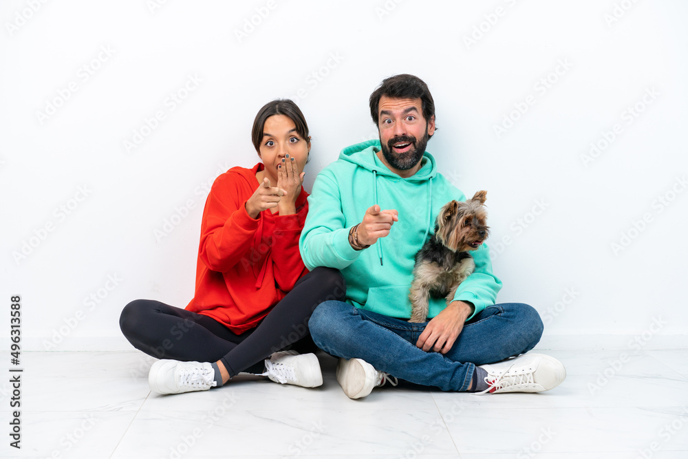 Young caucasian couple sitting on the floor with their pet isolated on white background pointing with finger at someone and laughing Young caucasian couple sitting on the floor with their pet isolated on white background pointing with finger at someone and laughing