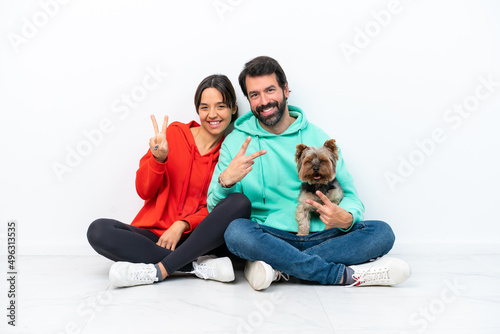 Young caucasian couple sitting on the floor with their pet isolated on white background smiling and showing victory sign