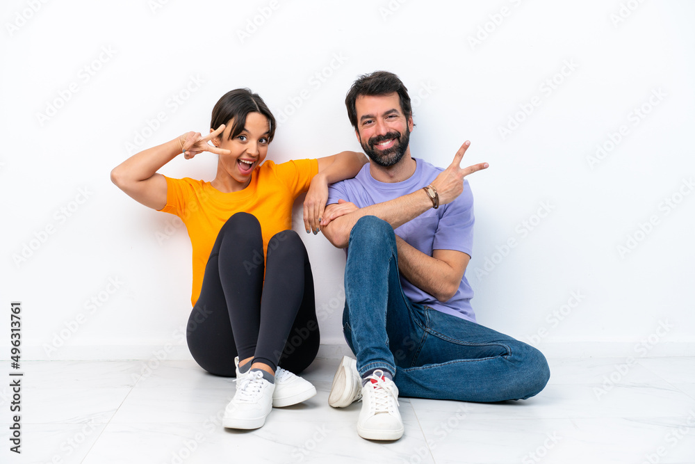 Young caucasian couple sitting on the floor isolated on white background smiling and showing victory sign Young caucasian couple sitting on the floor isolated on white background smiling and showing victory sign