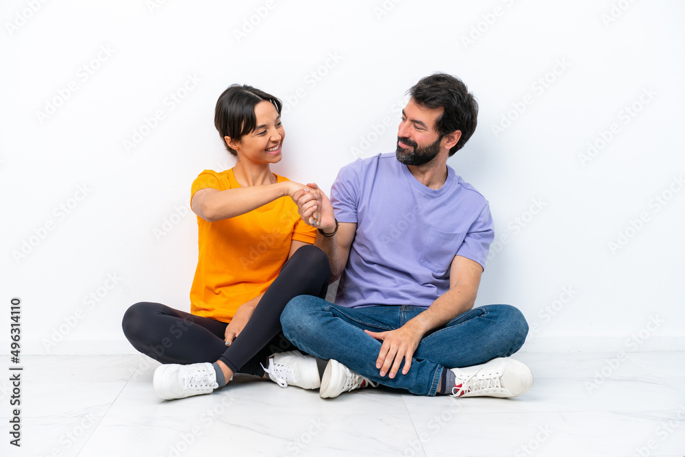 Young caucasian couple sitting on the floor isolated on white background handshaking after good deal Young caucasian couple sitting on the floor isolated on white background handshaking after good deal