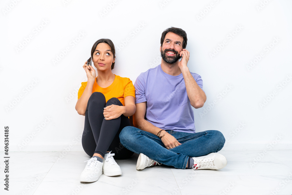 Young caucasian couple sitting on the floor isolated on white background talking to mobile Young caucasian couple sitting on the floor isolated on white background talking to mobile