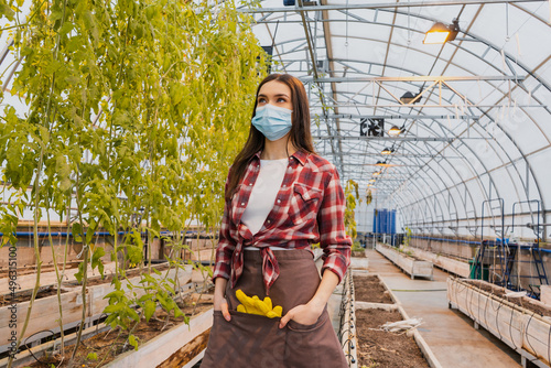 Young farmer in medical mask and apron looking at plants in greenhouse.
