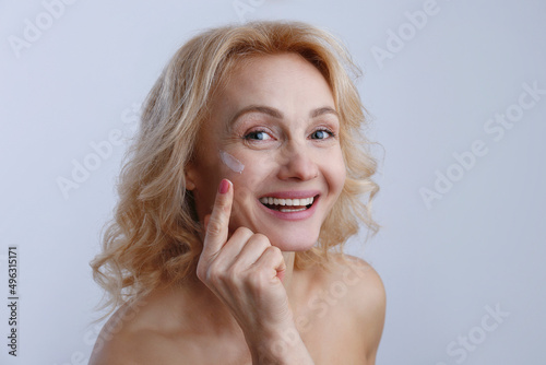 Portrait of adult woman with perfectly clean face skin applying moisturizing cream. Mid aged female with wavy blonde hair smiling performing daily skincare routine. Close up  copy space background.